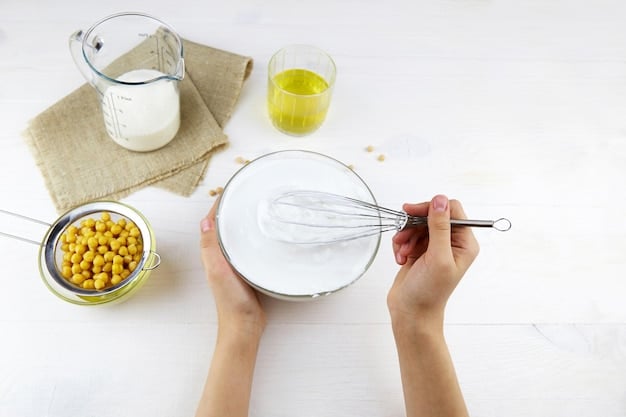 A close-up shot of hands mixing baking soda and vinegar in a glass bowl, with a wooden spoon stirring the mixture. The bowl is placed on a wooden countertop. The background is blurred, showcasing other natural cleaning ingredients such as lemons and essential oils.