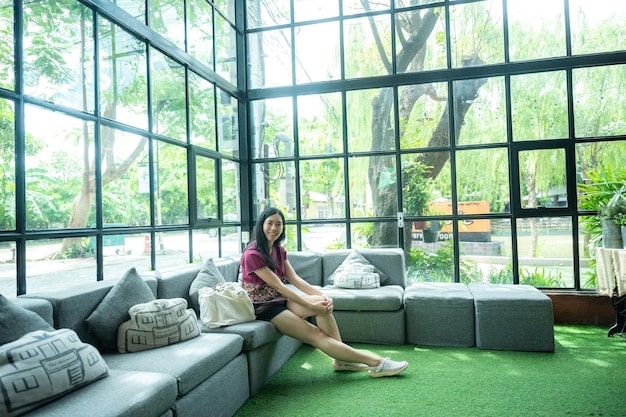 A brightly lit living room with large windows showcasing a lush green outdoor space. The focus is on energy efficiency, with LED lighting fixtures, a smart thermostat on the wall, and thick curtains drawn to insulate the room. The furniture is minimalist and eco-friendly, made from recycled materials.