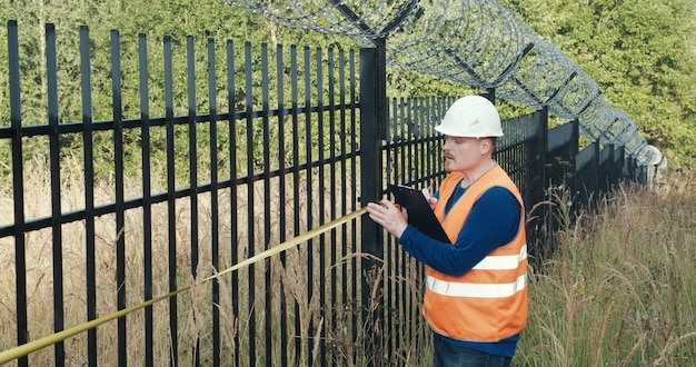 A close-up shot of a landowner inspecting a newly installed fence designed to protect a riparian area from overgrazing, with a clear stream and lush vegetation visible in the background. The landowner is wearing work gloves and appears to be carefully checking the fence's integrity.